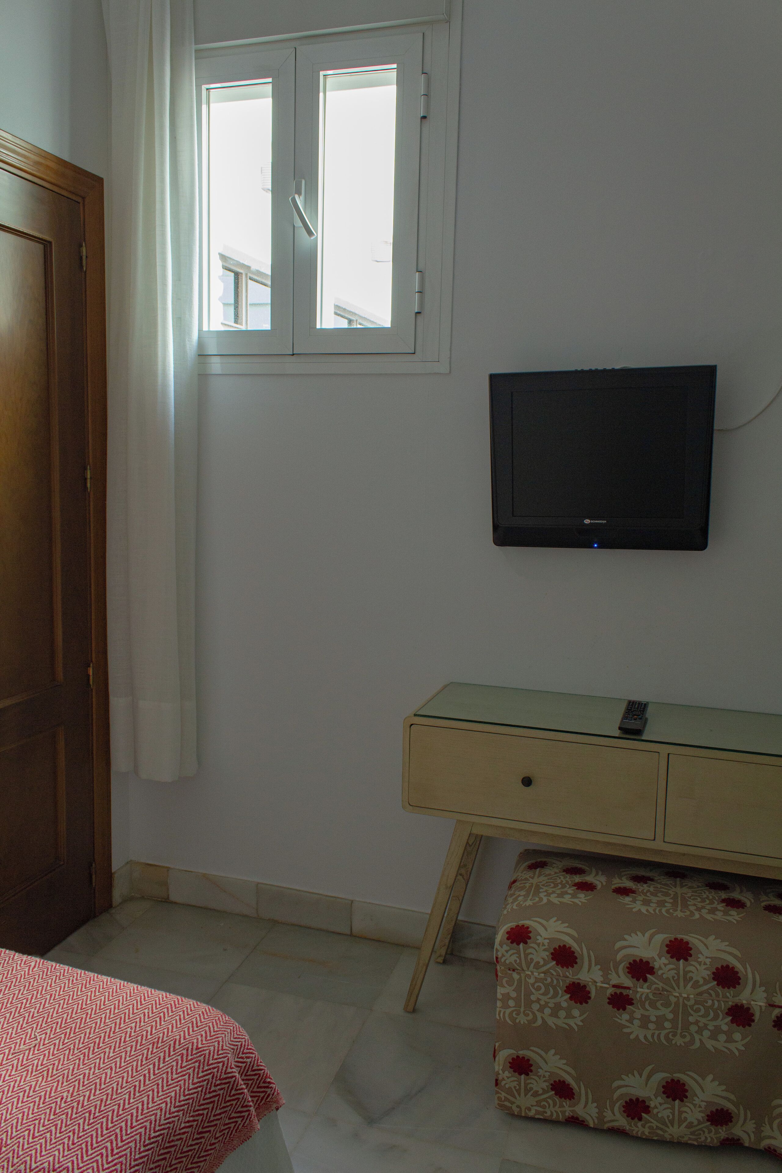A room with a bed, a wooden door, a window with curtains, a television mounted on the wall, and a small table with a remote control.