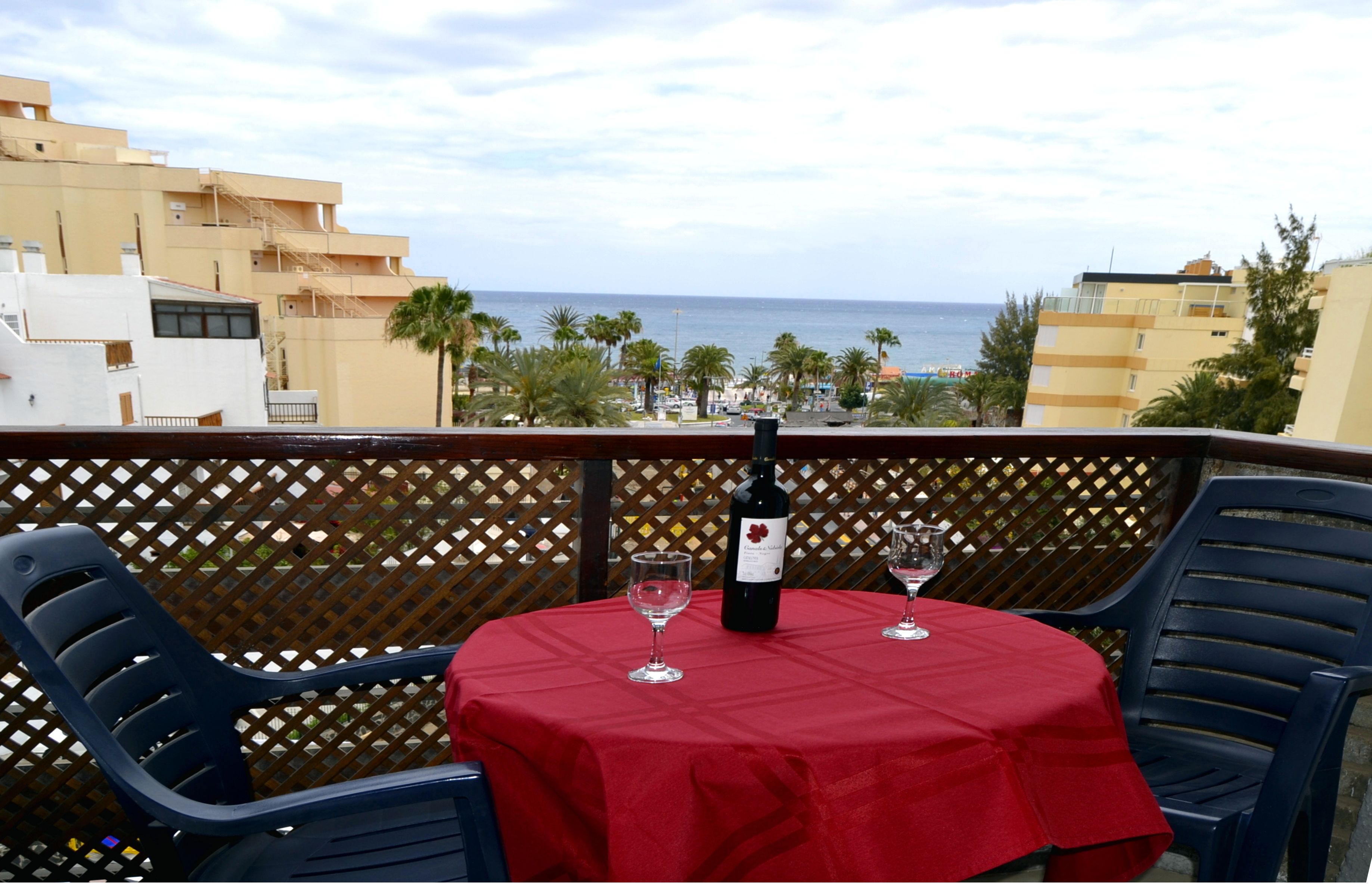 Un balcon avec une nappe rouge, deux chaises, une bouteille de vin et deux verres à vin. Le balcon donne sur une plage et des palmiers.