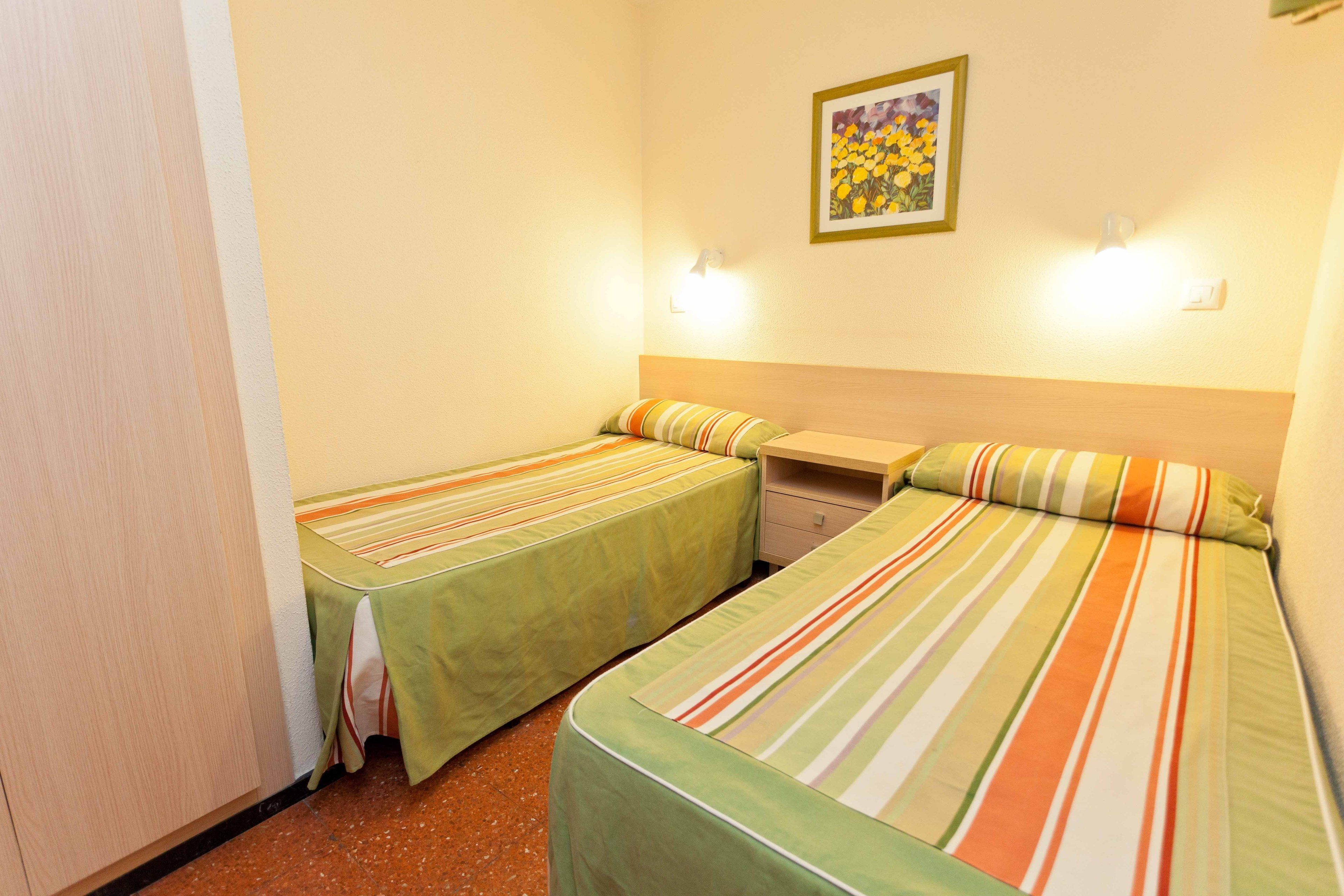 Ein kleines, sauberes Zimmer mit zwei Einzelbetten, jeweils mit gestreifter Bettwäsche, einem kleinen Nachttisch und einer an der Wand befestigten Lampe.