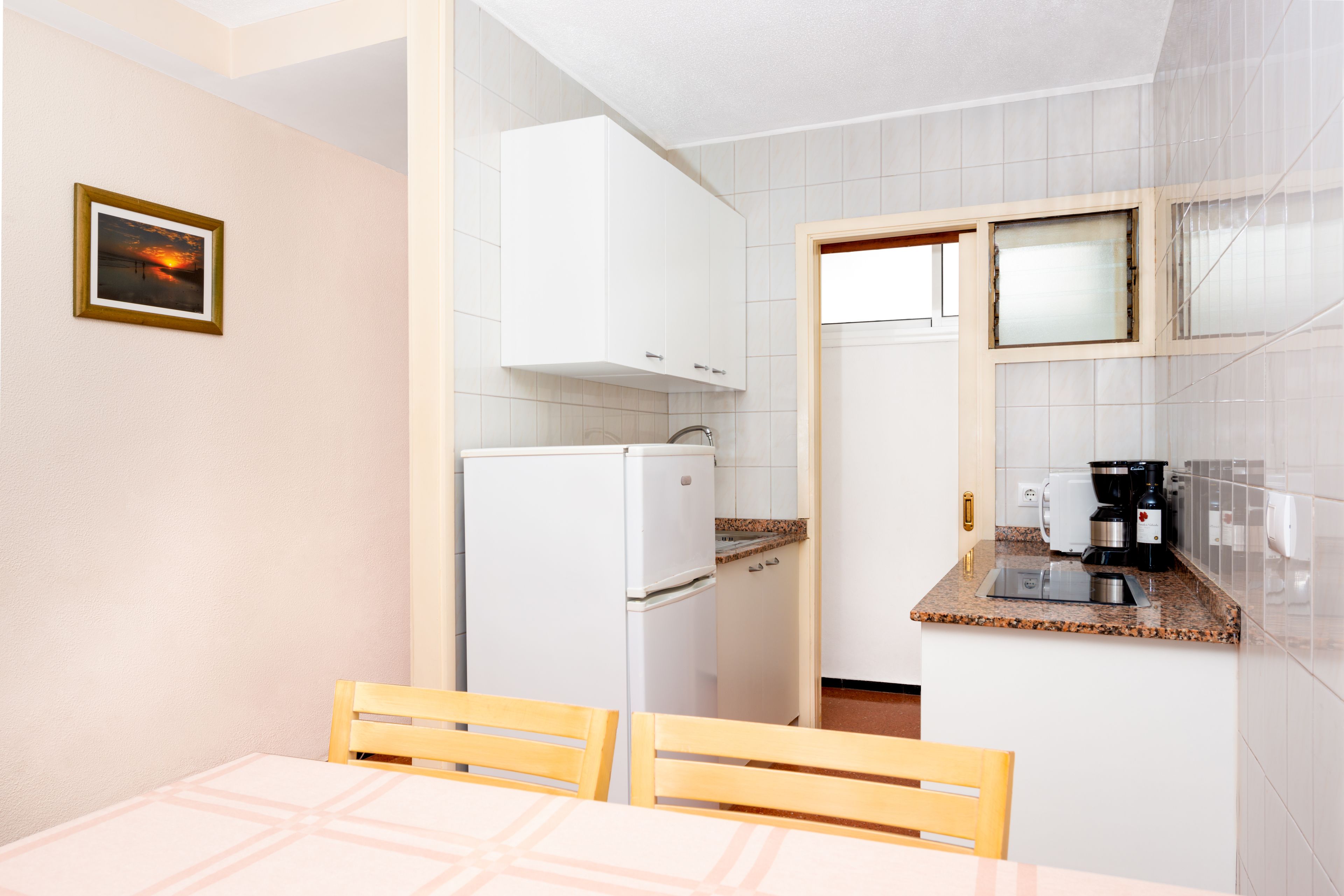 Una cocina compacta con armarios blancos, un refrigerador, una encimera con cafetera y una mesa de comedor con sillas.