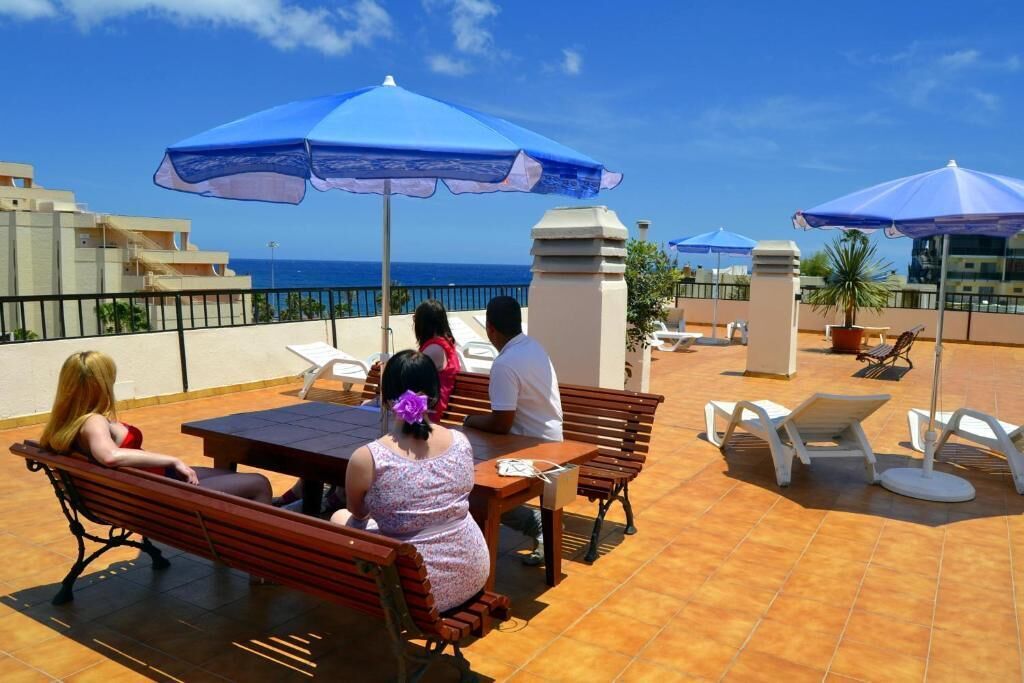 A terrace with wooden benches and tables, blue umbrellas, and a view of the sea.