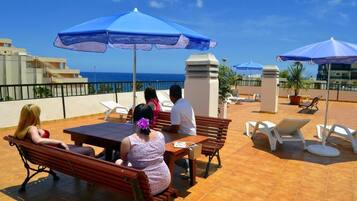 A terrace with wooden benches and tables, blue umbrellas, and a view of the sea.