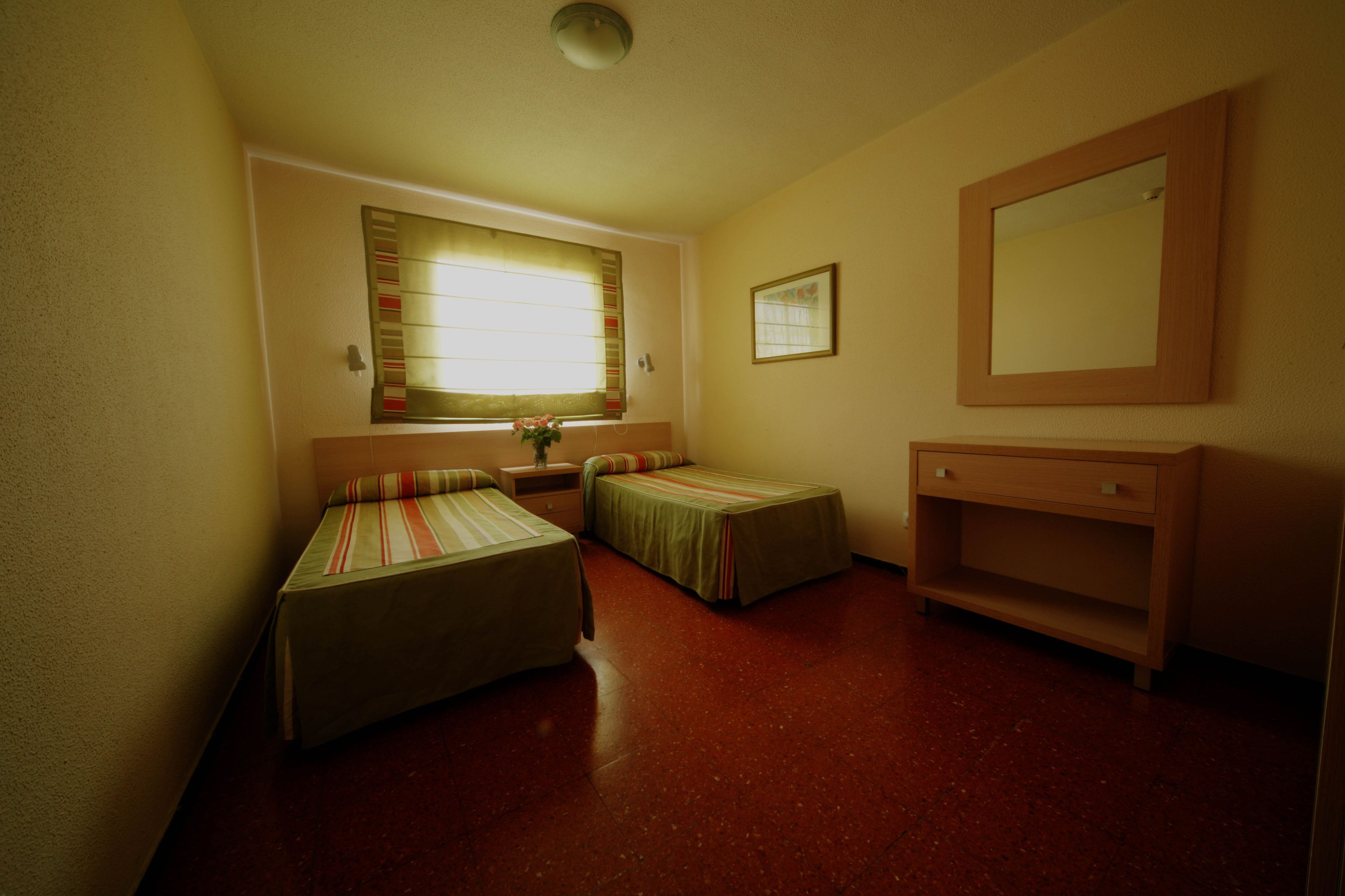 Une petite chambre compacte, équipée de deux lits simples, d’une table de chevet, d’un miroir et d’une fenêtre avec des stores.