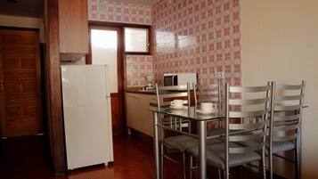 Cozinha compacta com geladeira branca, micro-ondas e cafeteira na bancada. Há duas cadeiras de metal e uma mesa de vidro. A cozinha possui uma porta de madeira e papel de parede estampado.