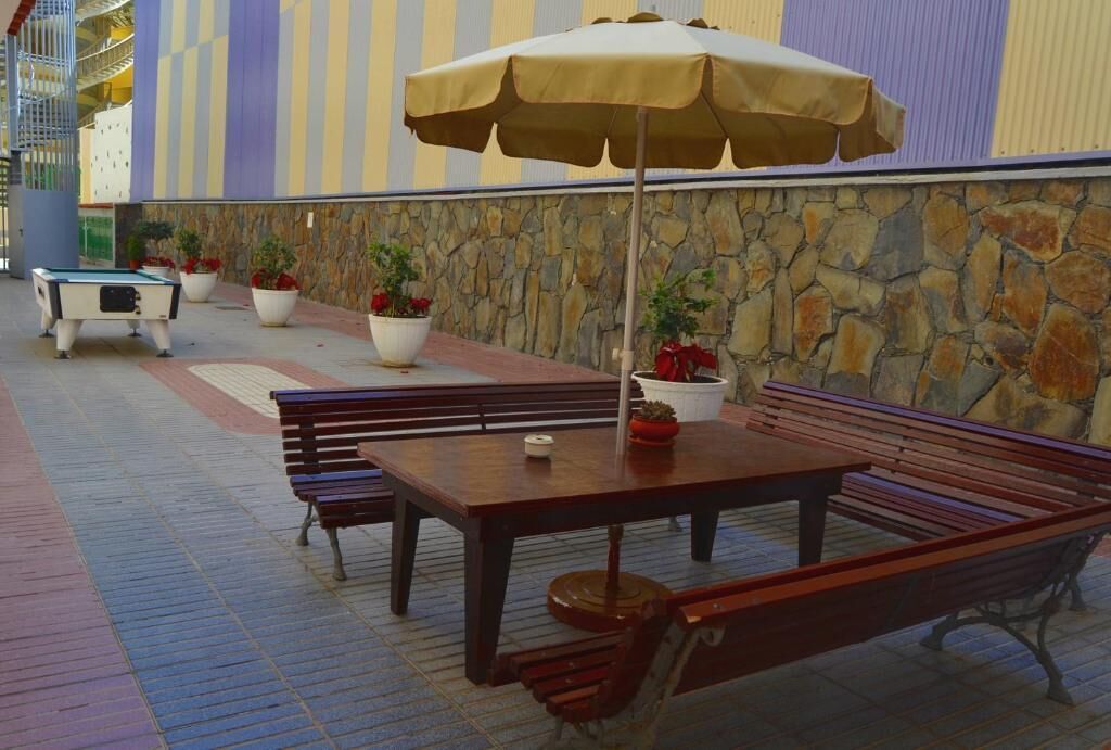 An outdoor seating area with wooden benches and a table under a large umbrella, surrounded by potted plants and a stone wall.