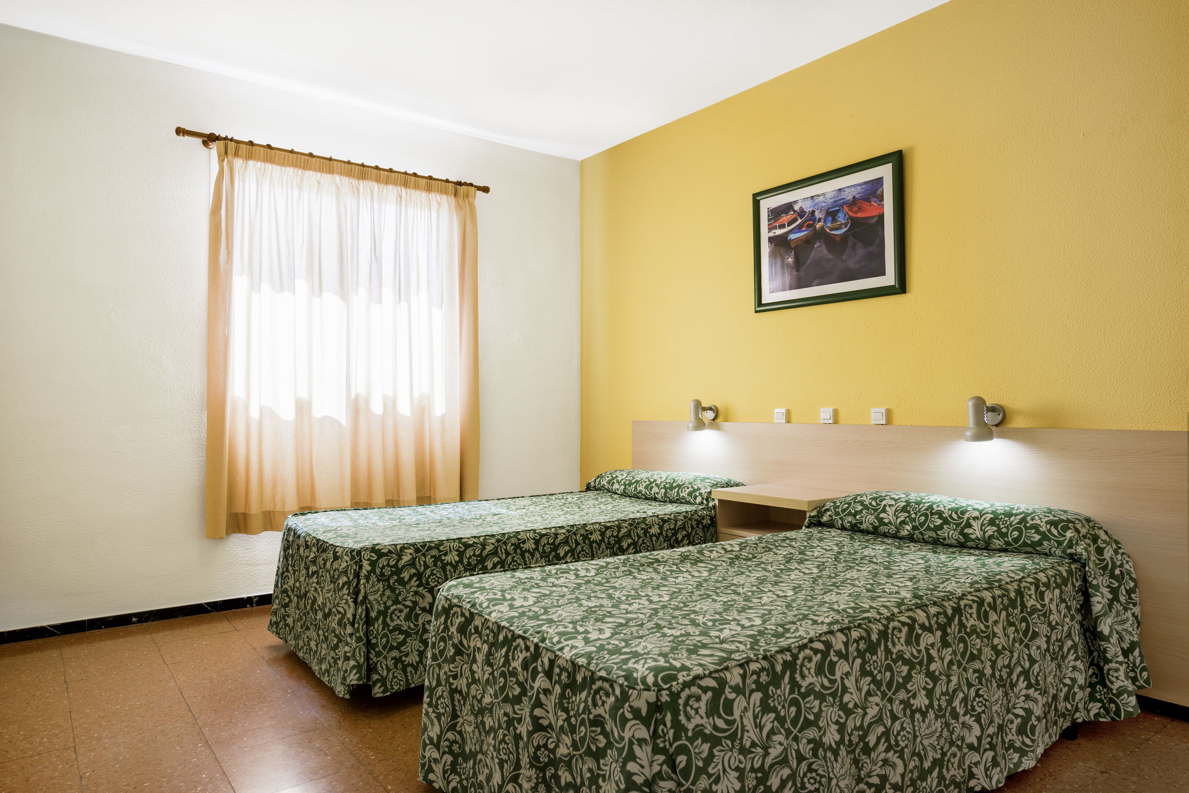A hotel room with two single beds, a window with sheer curtains, a yellow wall, and a framed picture above the bed.