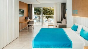 A hotel room with a large bed, a desk, a chair, a sofa, and a balcony with a view of trees and a building.