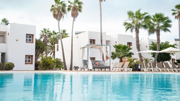 A swimming pool with lounge chairs and umbrellas, surrounded by palm trees and white buildings.