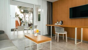 A modern hotel room with a wooden accent wall, a flat-screen TV, a dining area with a table and chairs, and a sofa.