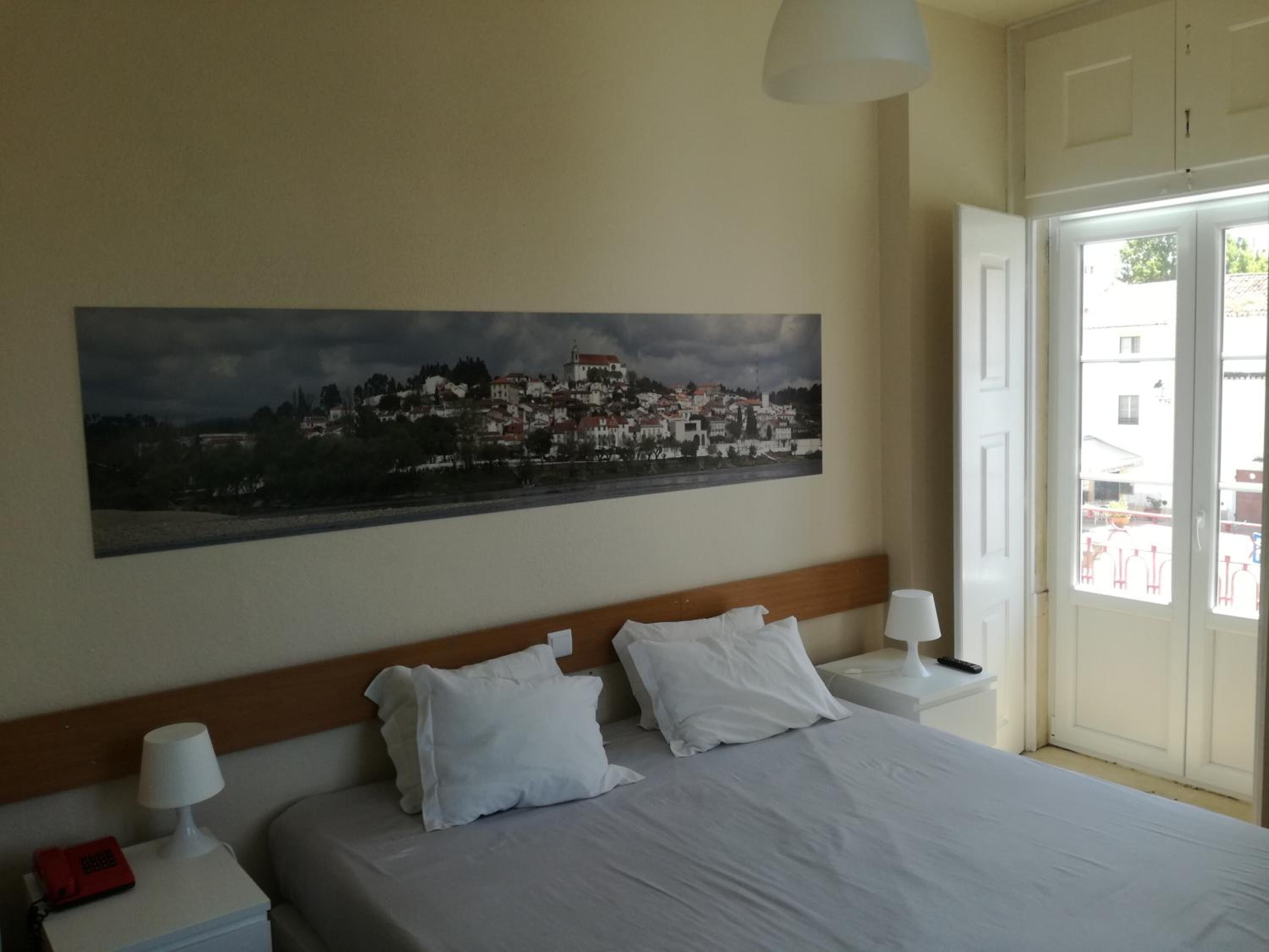 Een hotelkamer met een bed, twee nachtkastjes, een lamp, een schilderij van een stadsgezicht en een deur die uitkomt op een buitenruimte.