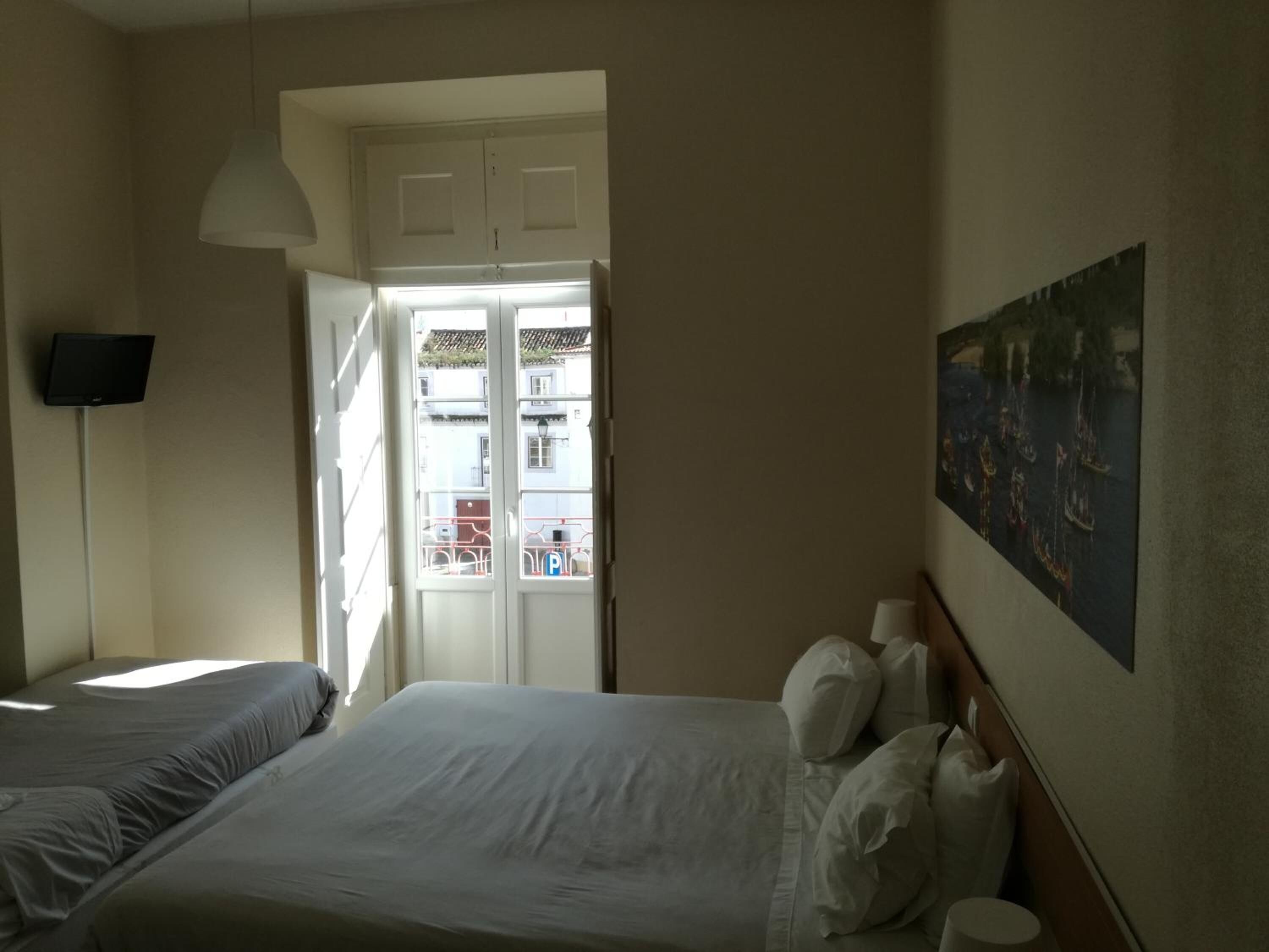A hotel room with two beds, a flat-screen TV, a painting on the wall, and a view of the street through an open door.