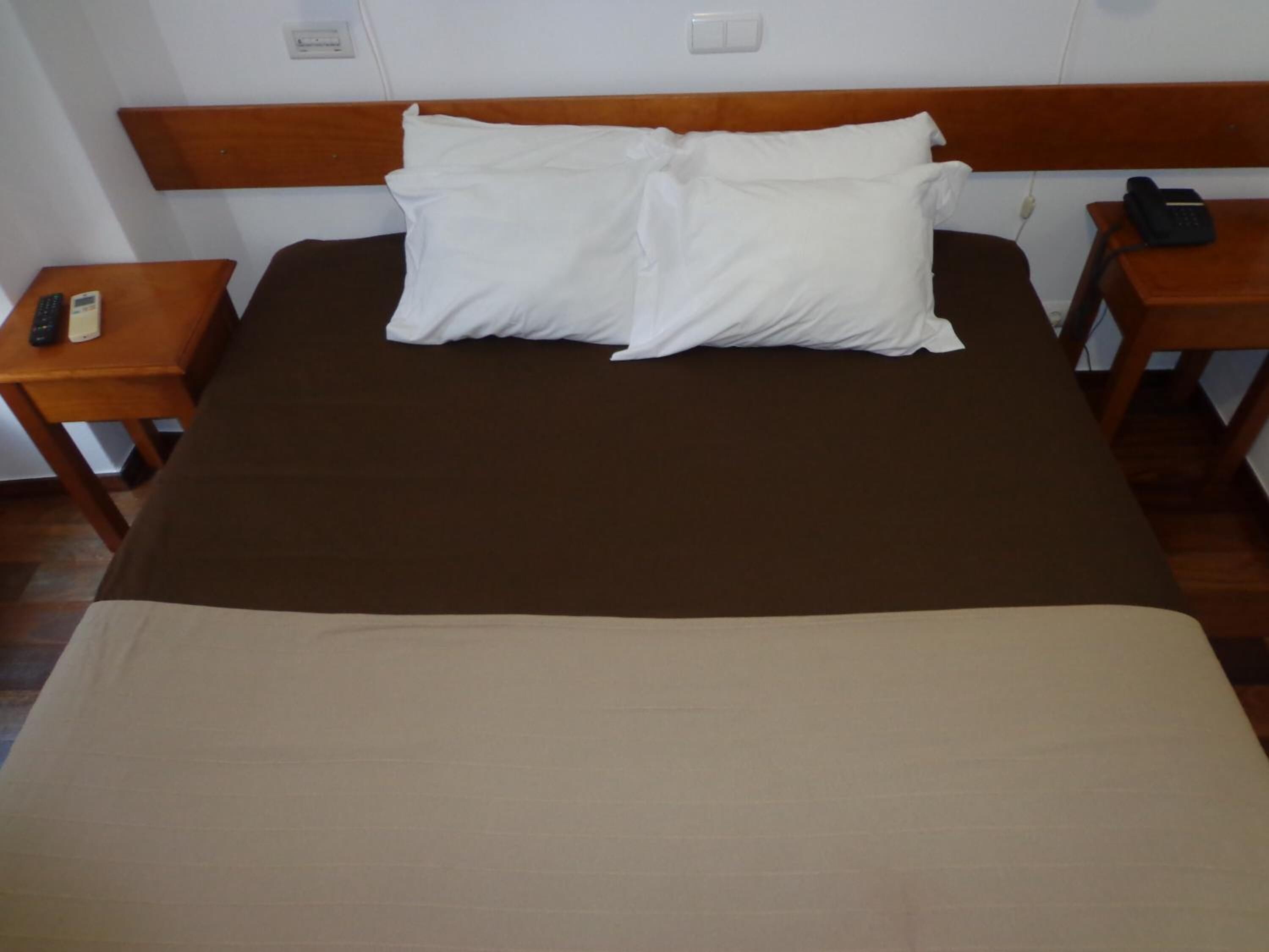 A bed with a brown bedspread and two white pillows, flanked by wooden nightstands with a remote control and a phone on one.