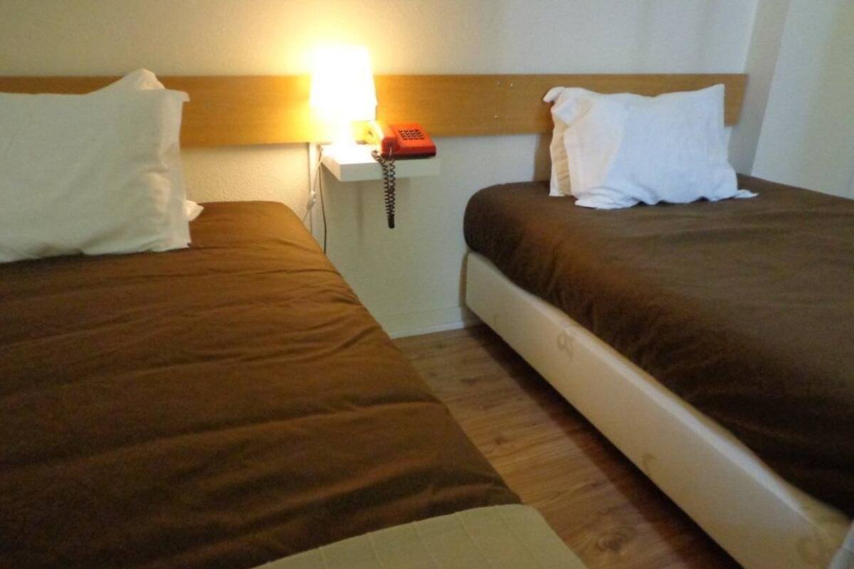 A hotel room with two beds, each with a pillow and a blanket. There is a bedside table with a lamp and a telephone.