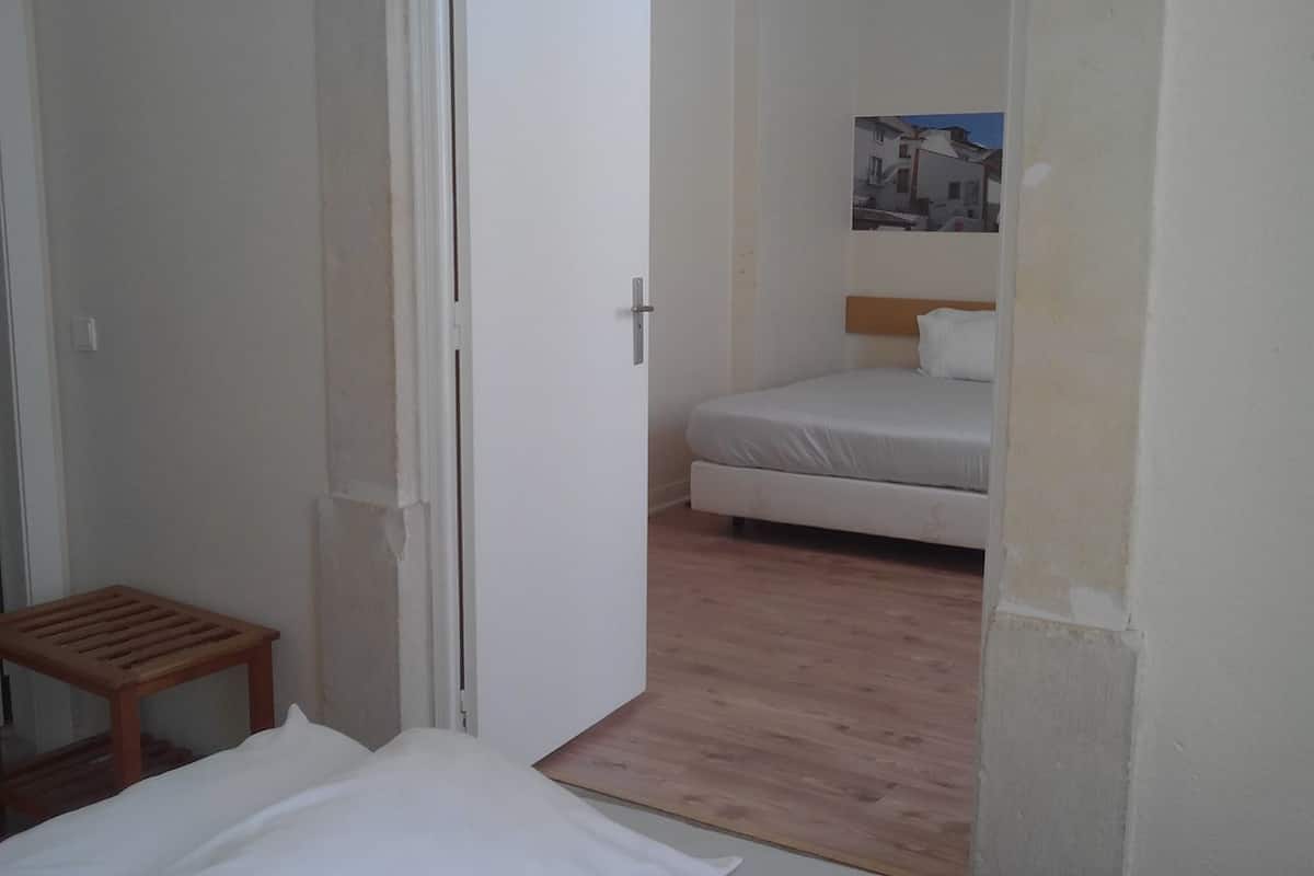 A small, clean room with a bed, a wooden stool, and a door leading to another room.