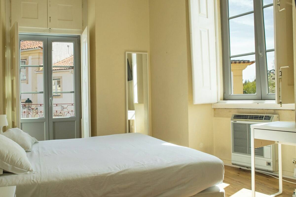 A hotel room with a bed, a desk, a window, and an air conditioning unit.