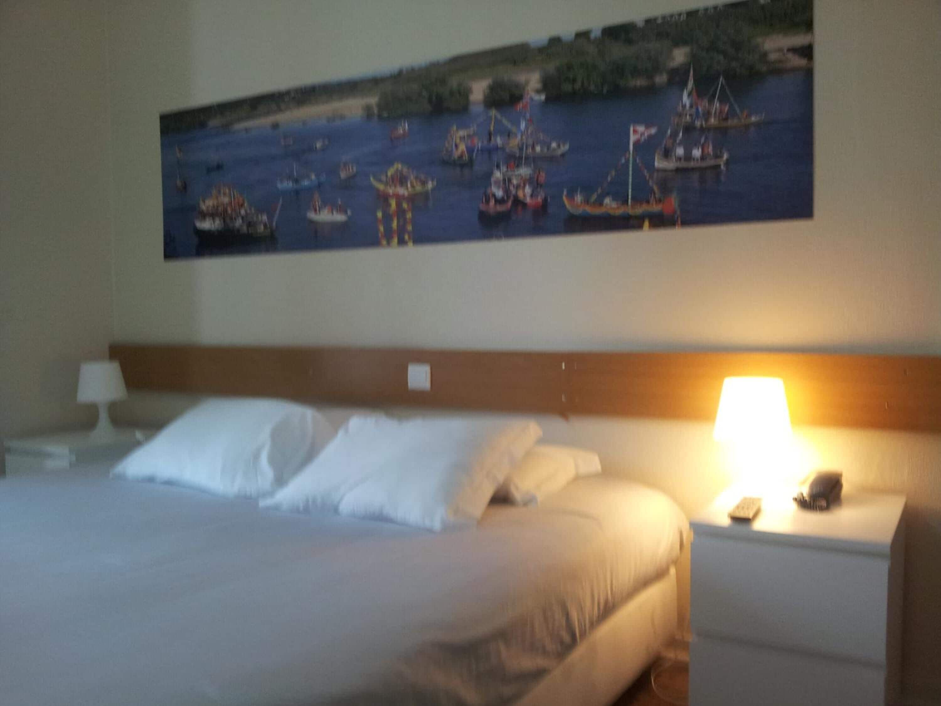 Quarto de hotel com cama, mesa de cabeceira, luminária e um quadro de barcos na parede.