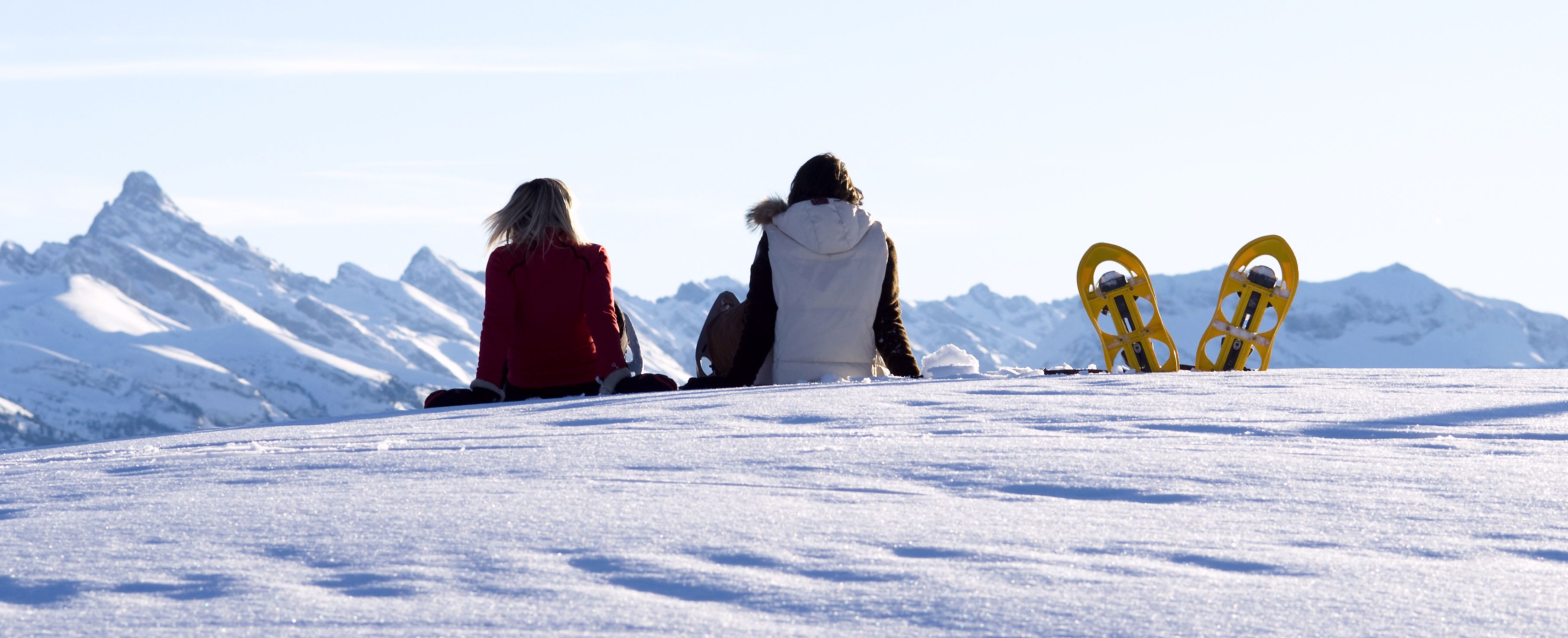 Two people sitting on a snowy slope with snowboards, facing a mountain range.