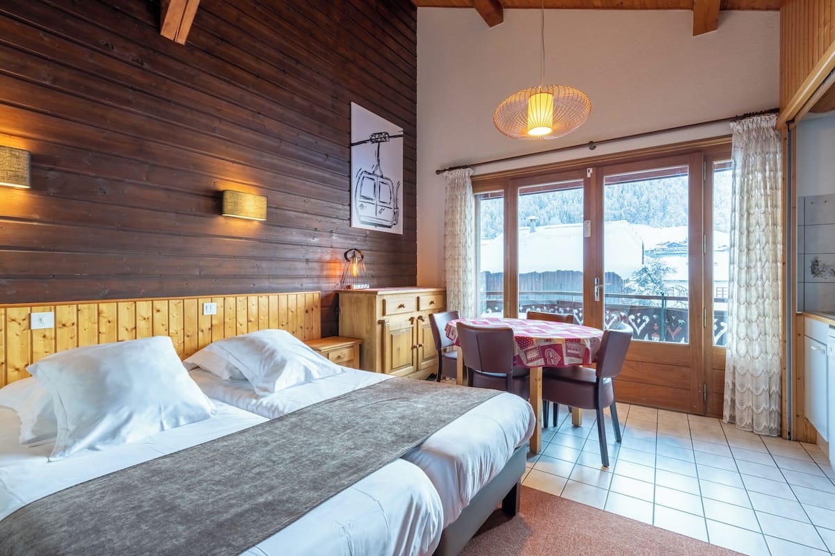 Una habitación acogedora con cabecera de madera, cama con ropa de cama blanca, cómoda de madera, mesa de comedor con sillas y un ventanal con vista a un paisaje nevado.