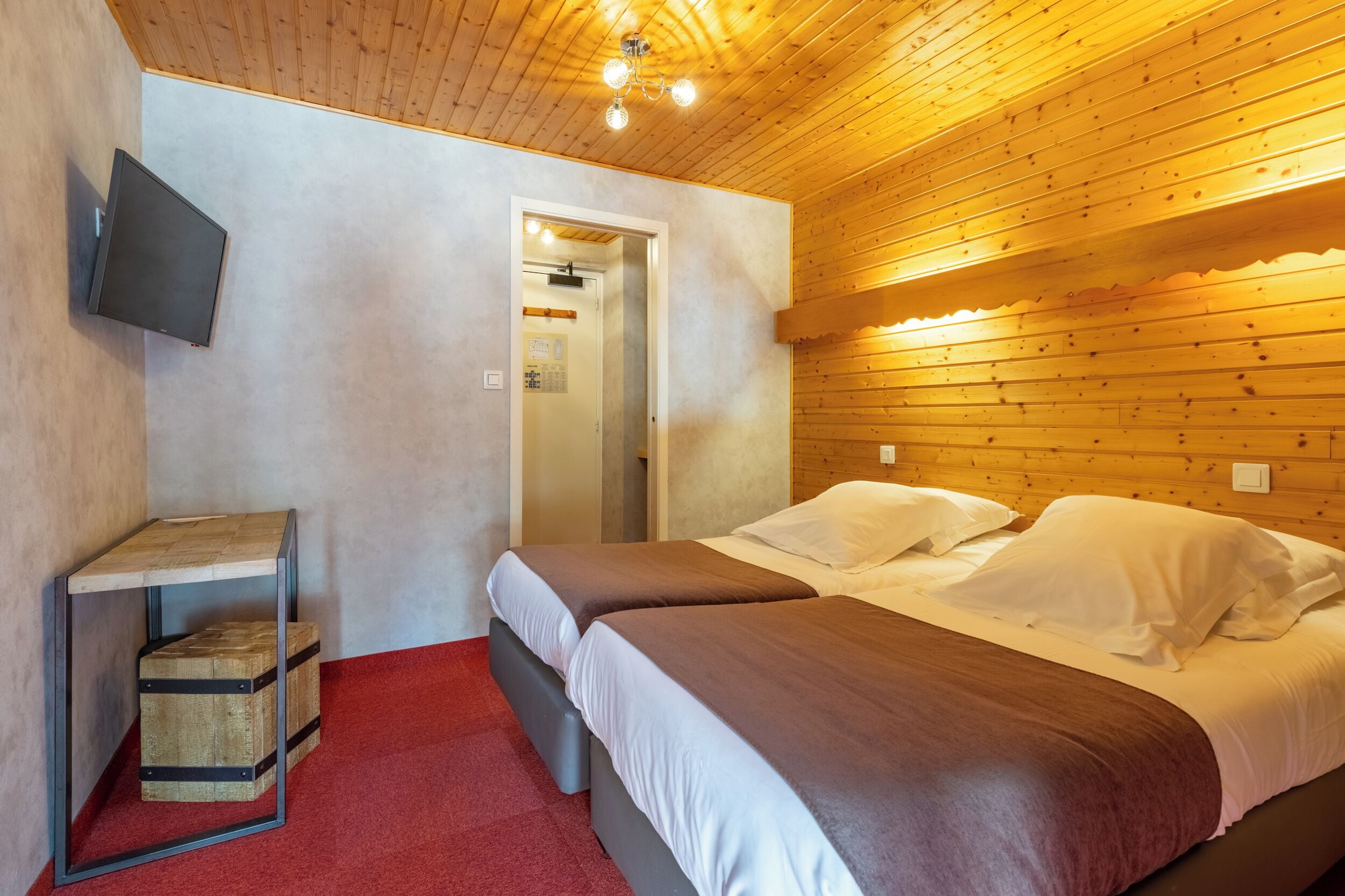 A room with two beds, a TV mounted on the wall, a wooden ceiling, and a red carpet.