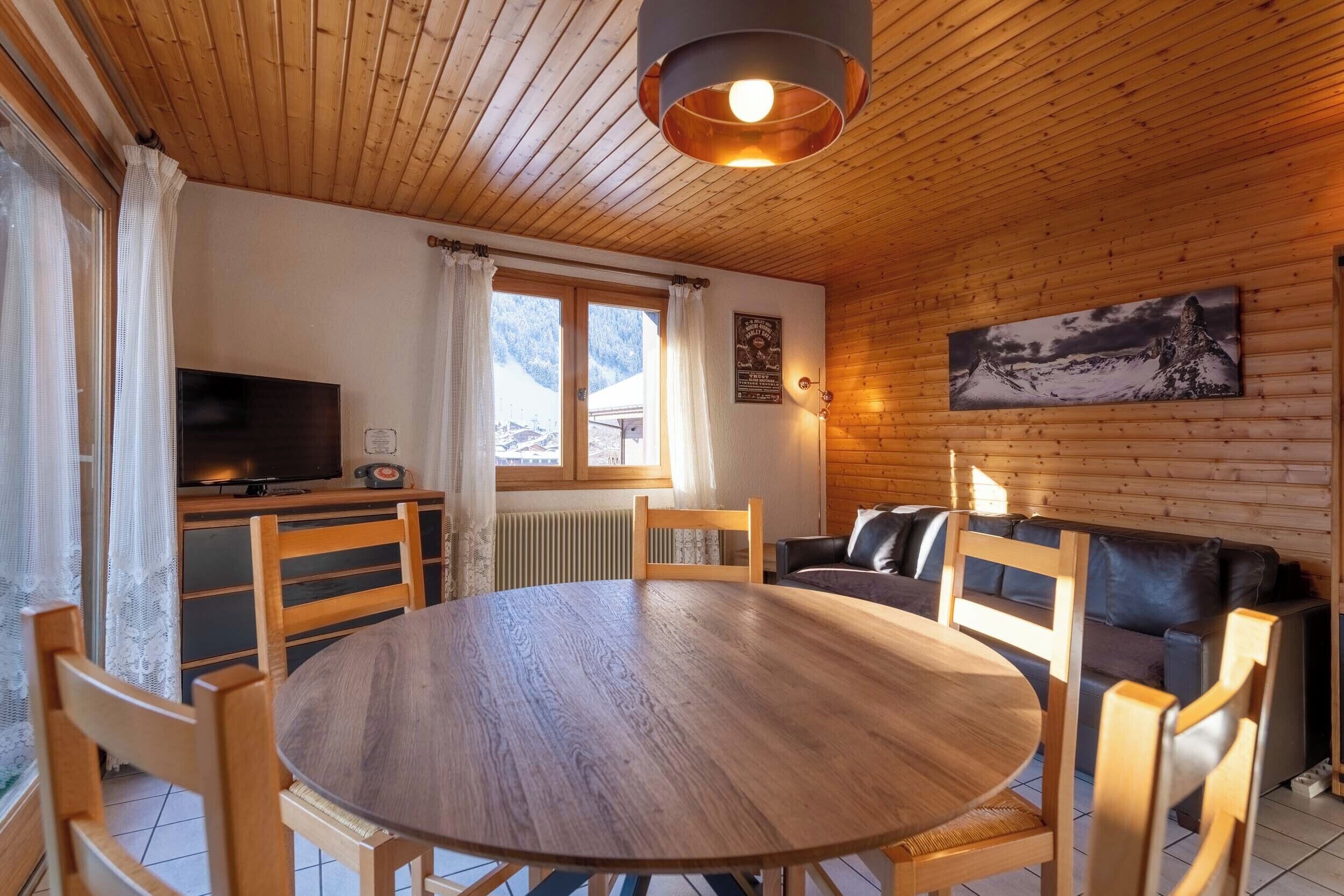 Ein gemütliches Zimmer mit Holzbalkendecke, einem runden Holztisch mit Stühlen, einem Sofa, einem Fernseher und einem Fenster mit Vorhängen.