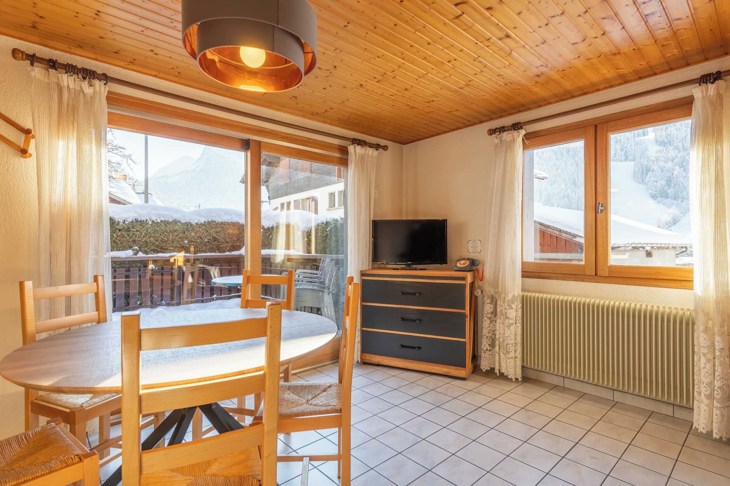 Une pièce chaleureuse avec un plafond en bois, une table à manger avec des chaises, un téléviseur posé sur un meuble et de grandes fenêtres avec des rideaux.