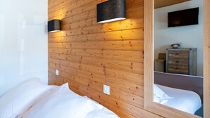 Een slaapkamer met houten paneelwanden, een bed met witte lakens, twee zwarte wandlampen en een spiegel die een dressoir en een televisie weerspiegelt.
