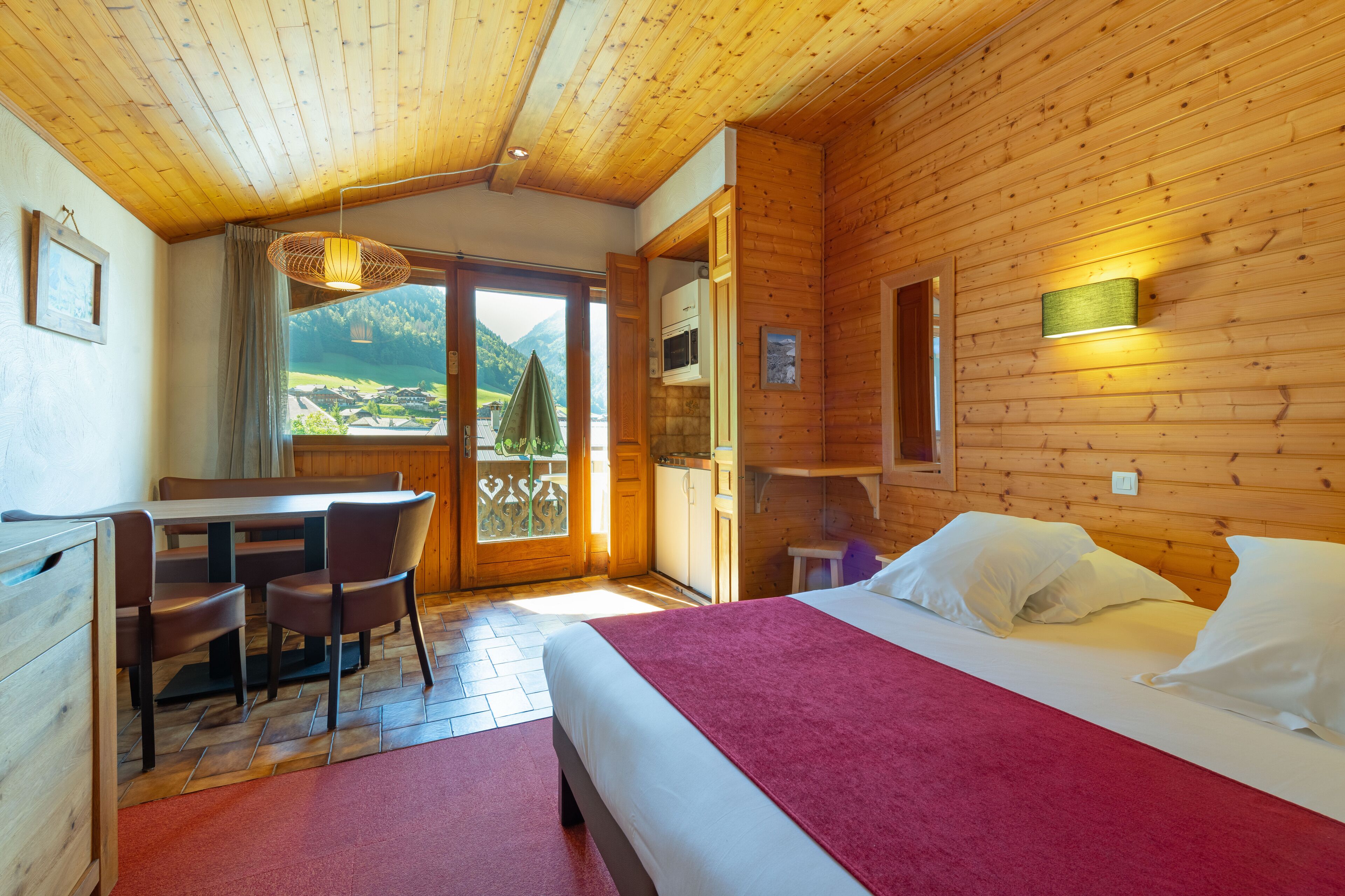 A wooden cabin room with a bed, a table with chairs, a kitchen area, and a view of the outdoors.