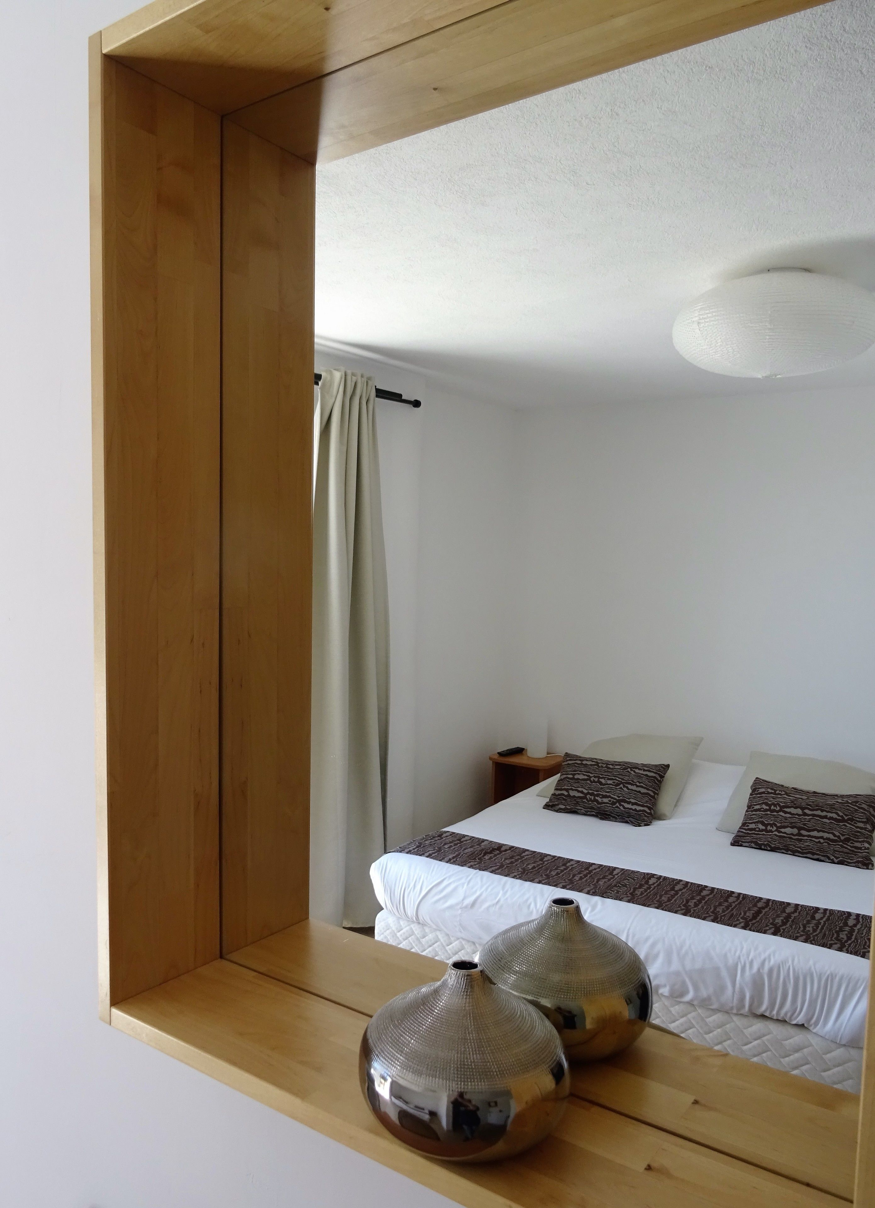 Une chambre à coucher avec une tête de lit en bois, un lit avec une literie blanche et des oreillers à motifs, une table de chevet avec une lampe, et deux vases décoratifs sur une étagère en bois.