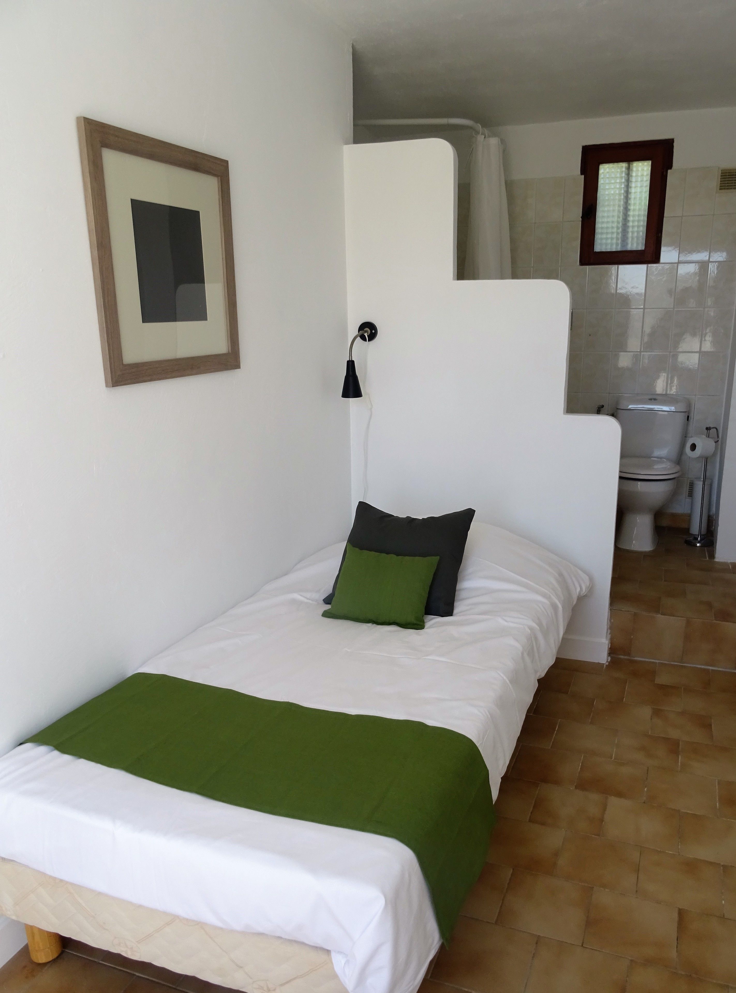 A small, single-bed room with a green and white bedspread, a framed picture on the wall, and a bathroom visible through an open door.