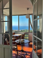 Un balcón abierto con una mesa dispuesta para el desayuno, una lámpara colgante y vistas al mar y al paisaje distante.