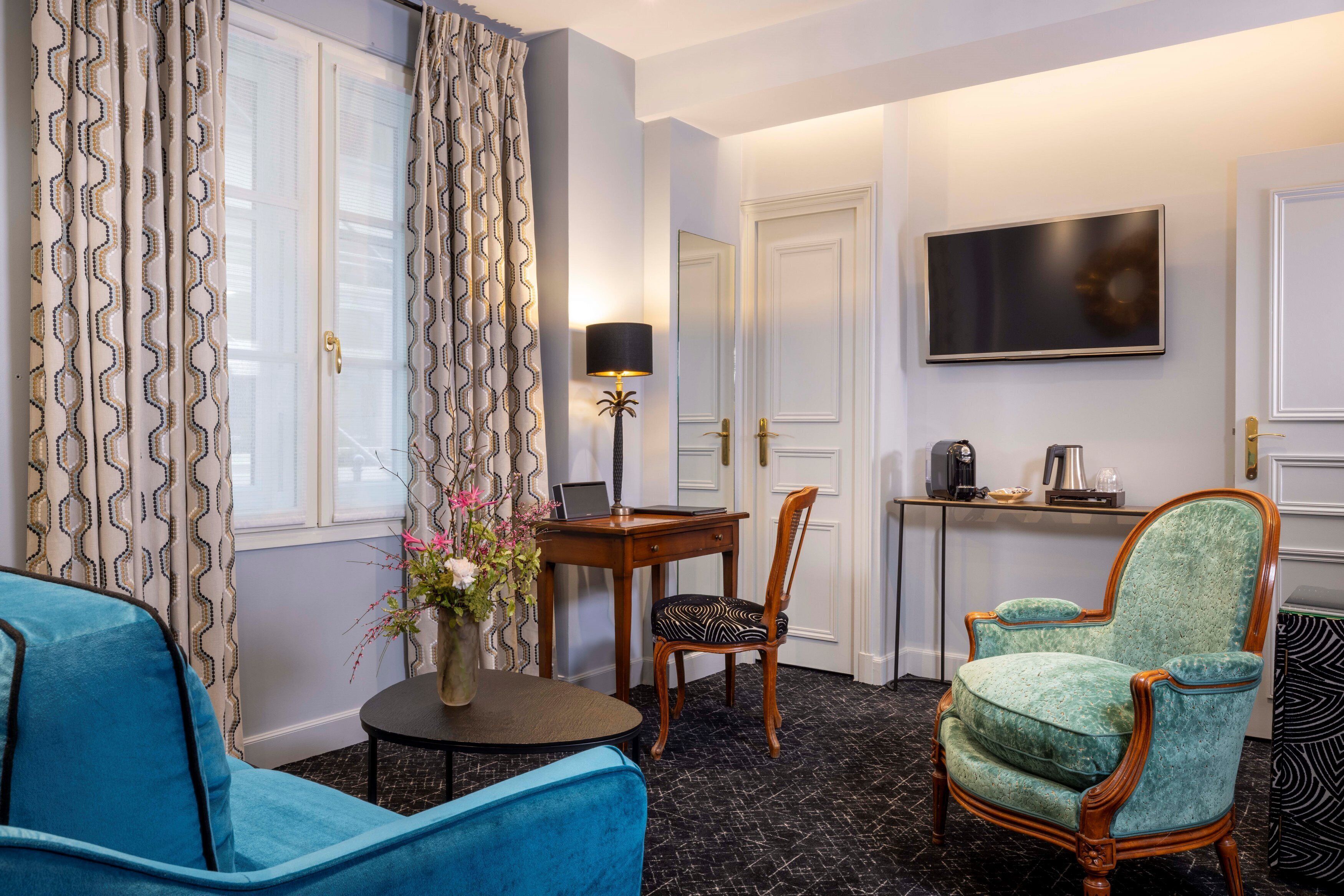Une chambre d’hôtel avec un fauteuil bleu, une chaise en bois, une petite table ronde avec une lampe, un téléviseur fixé au mur et un bureau avec une machine à café.