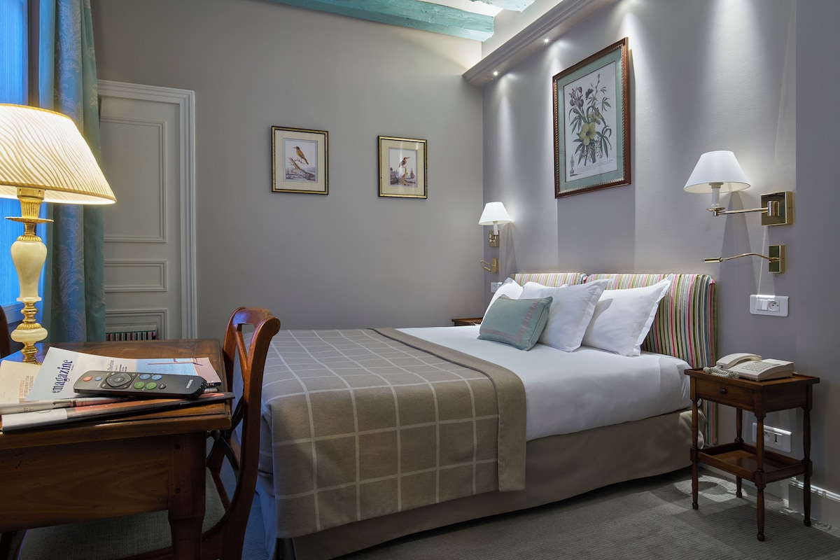 A neatly arranged hotel room with a bed, bedside lamps, a small table with a lamp, a chair, and a framed picture on the wall.