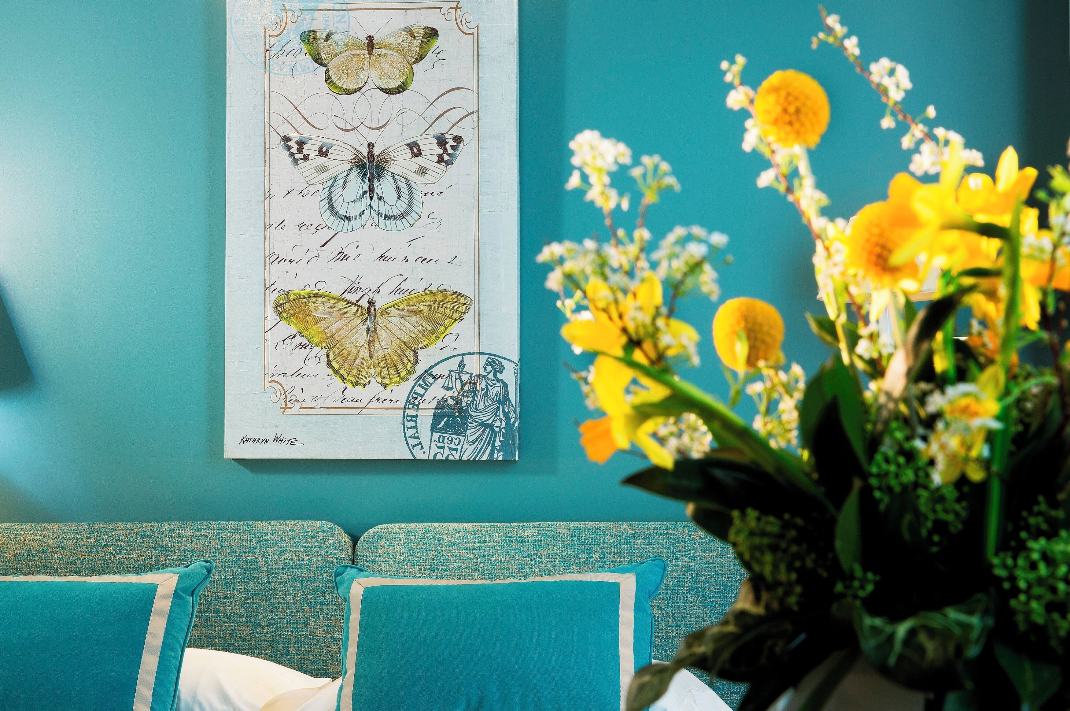 A teal-colored sofa with matching cushions, a framed botanical illustration on the wall, and a vase of yellow flowers.