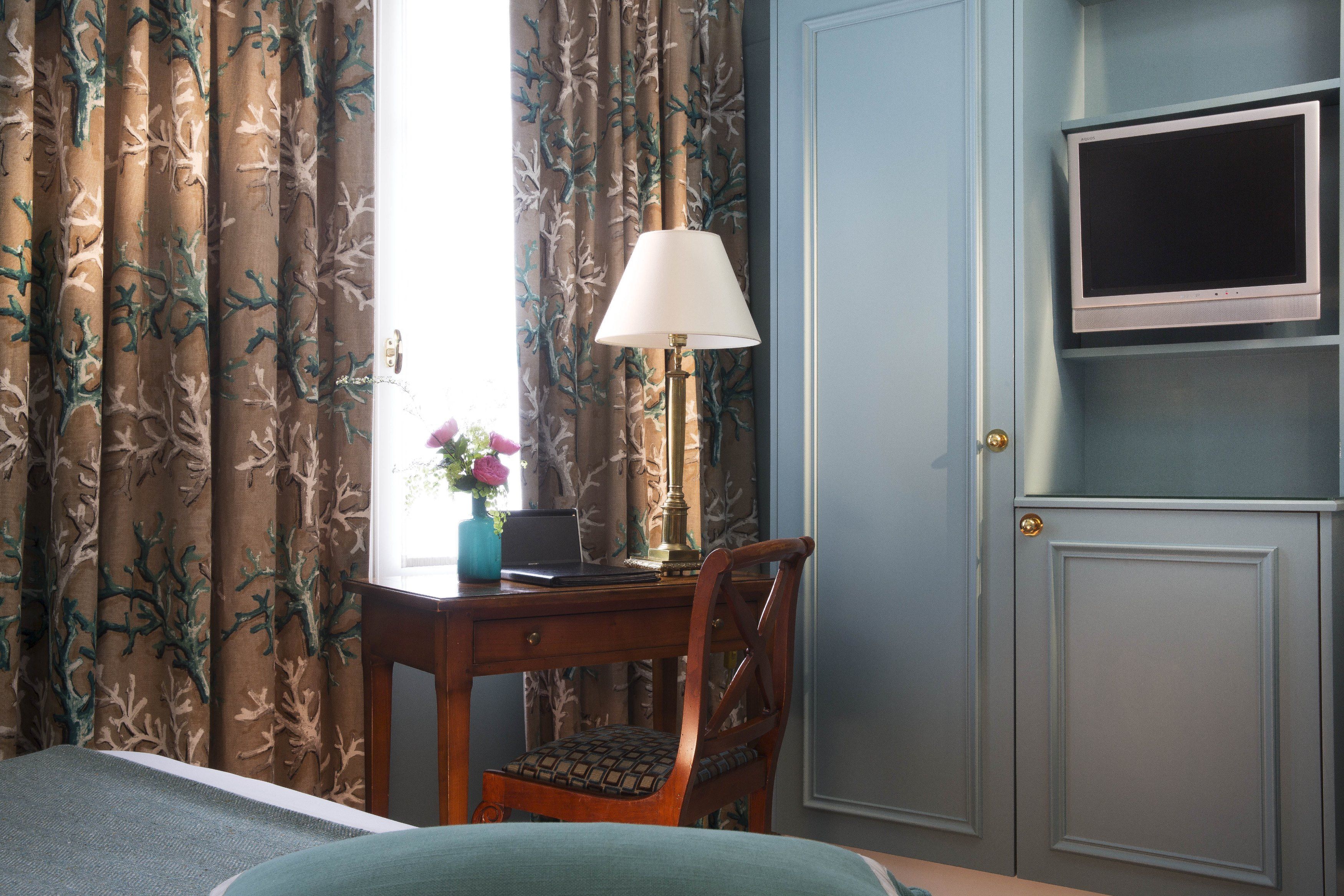 Habitación de hotel con cama, escritorio con lámpara, silla, televisor sobre un mueble bajo y cortinas con estampado de árboles.