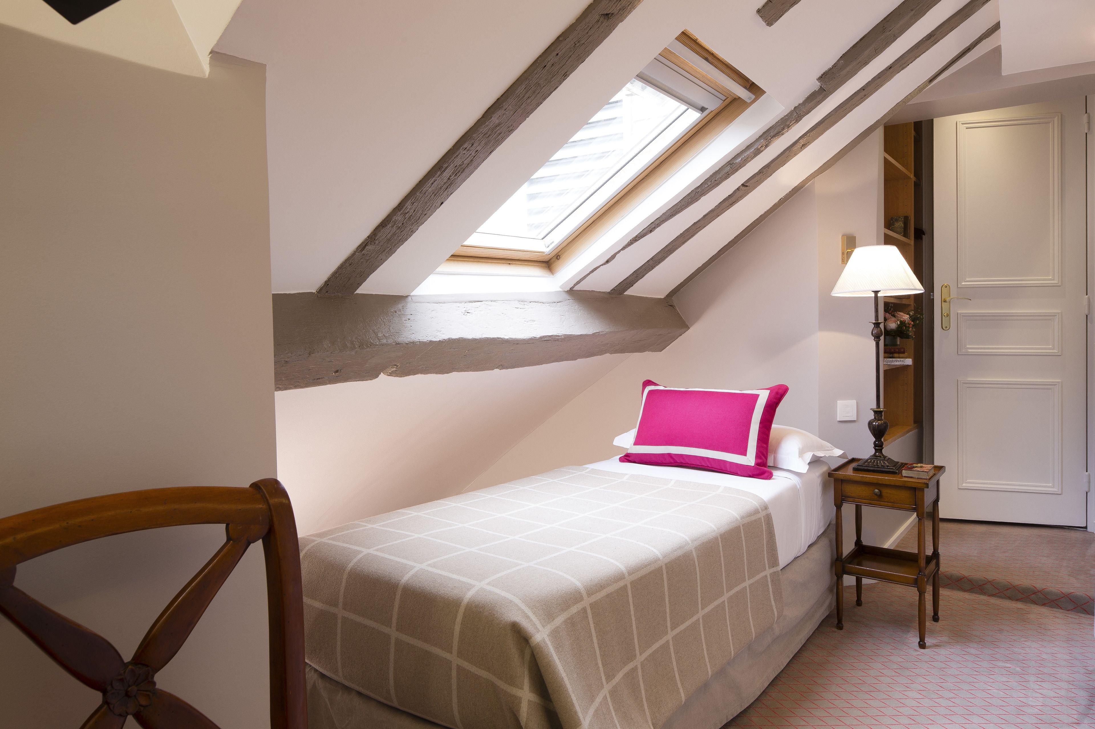 Un dormitorio con tragaluz, una cama con almohada rosa, una mesita de noche con lámpara y una silla de madera.