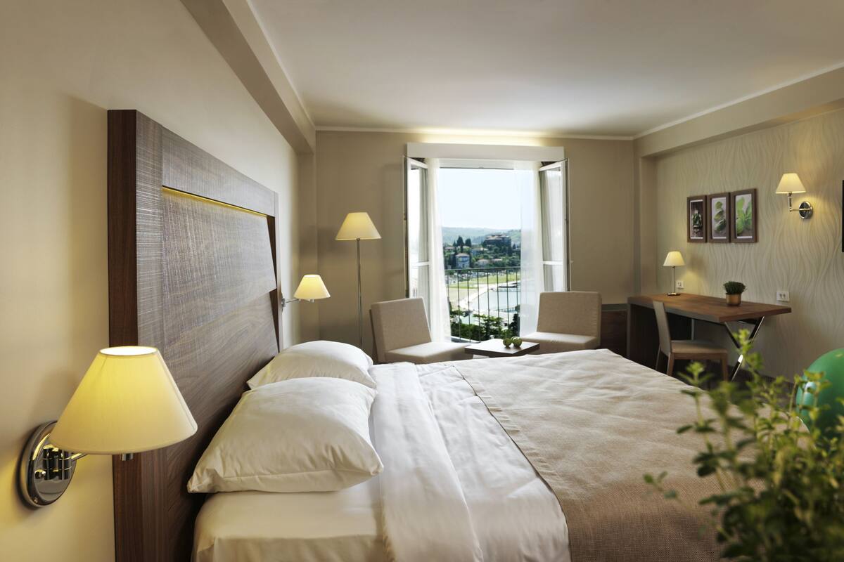 A hotel room with a bed, two chairs, a desk, a TV, and a balcony with a view.