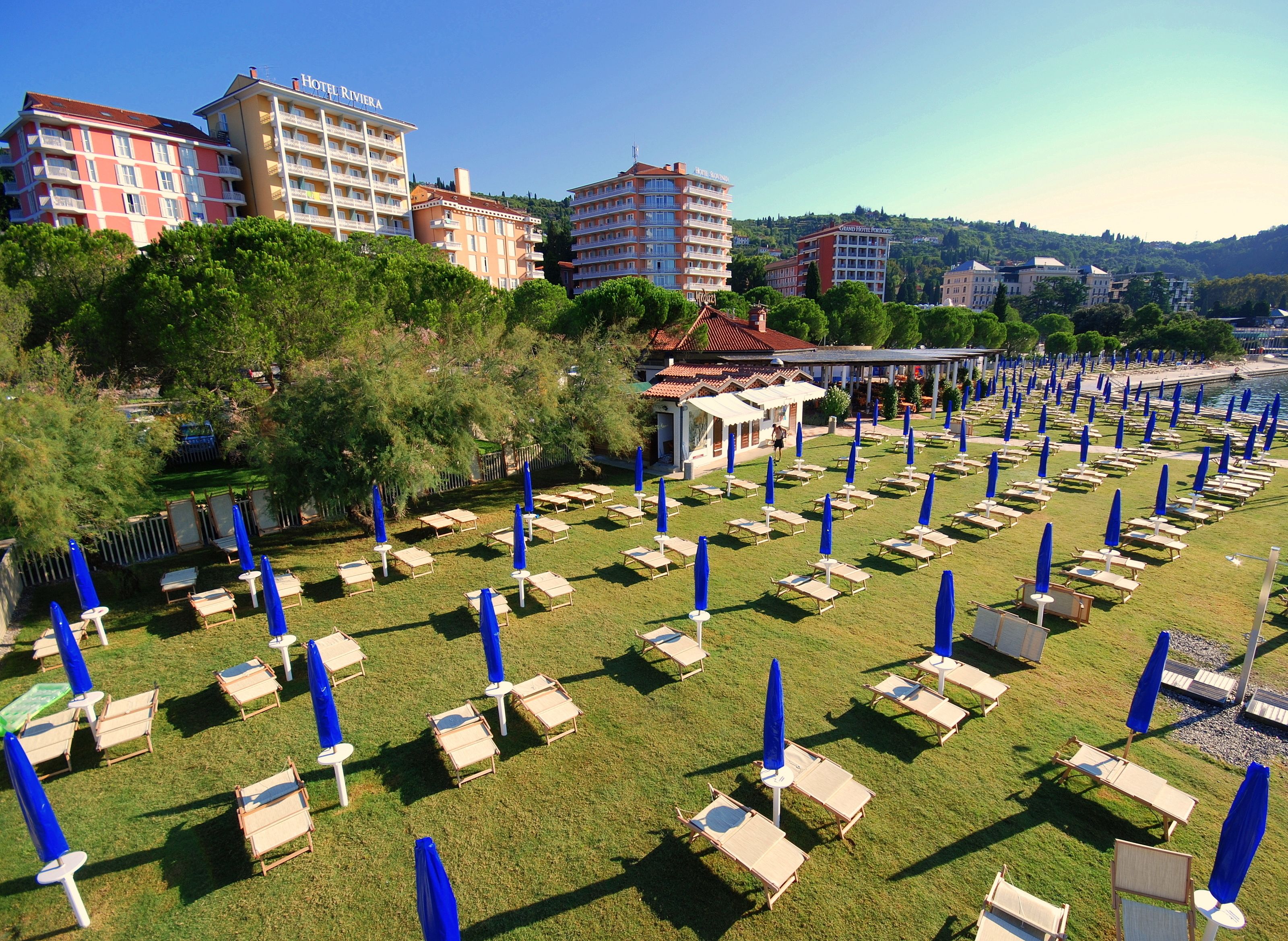 A hotel with multiple floors, outdoor seating areas with blue umbrellas, and a view of the sea.