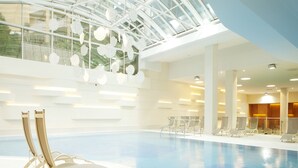 Une piscine intérieure à l’eau claire, avec des murs et un plafond blancs. On y trouve des chaises longues et un plafond en verre.