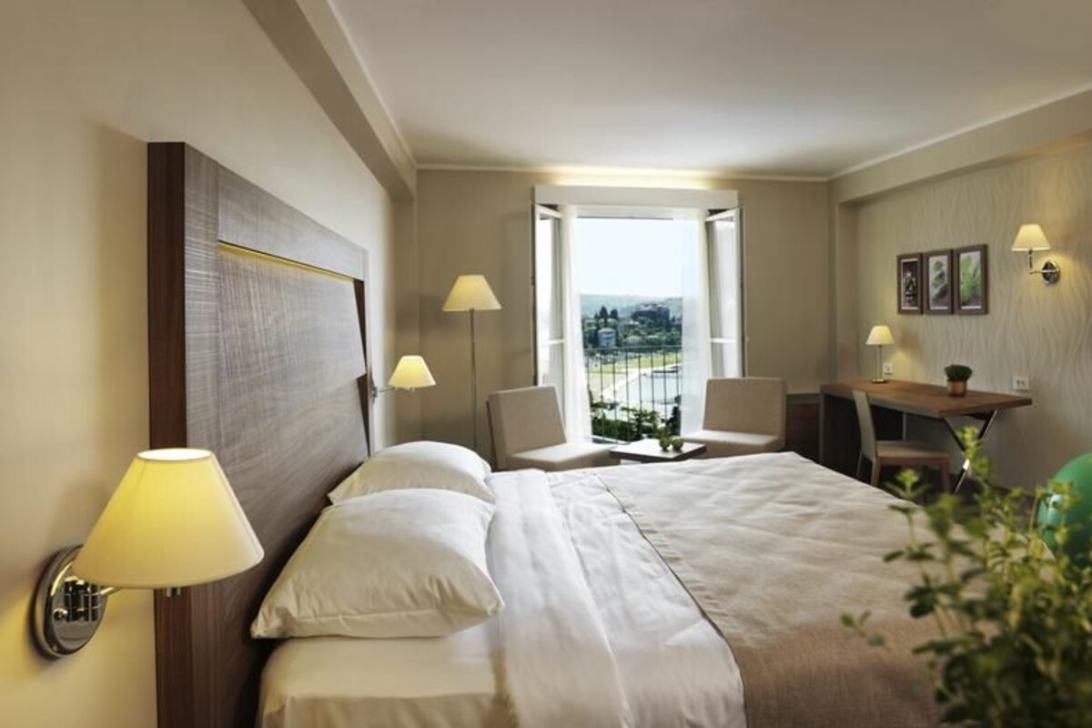 A hotel room with a bed, two chairs, a desk, a TV, and a balcony with a view.