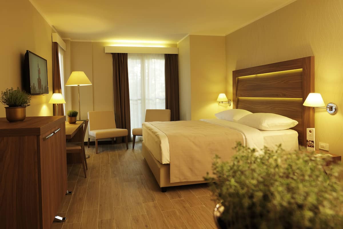 A hotel room with a bed, bedside lamps, a desk with a chair, a potted plant, and a view of the city skyline through the window.