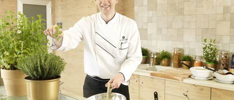 Un chef con uniforme blanco y gorro de cocinero, de pie en una cocina con alacenas de madera y salpicadero de azulejos. Se ven plantas en macetas, una vitrina de vidrio y diversos utensilios de cocina.