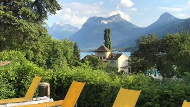 A terrace with yellow and white lounge chairs, a small table with a bottle, and a view of mountains and a lake. - Hotel Du Lac (Talloires-Montmin)