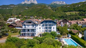 A large, multi-story building with balconies, a swimming pool, and a mountainous backdrop. - Hotel Du Lac (Talloires-Montmin)