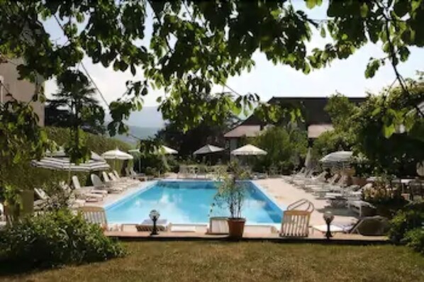 Una piscina con sillas de descanso y sombrillas, rodeada de vegetación y un edificio al fondo.