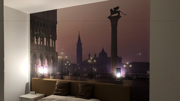 A hotel room with a large bed, two bedside tables, and a wall mural of a cityscape at dusk. - Hotel Angi (Fosso)