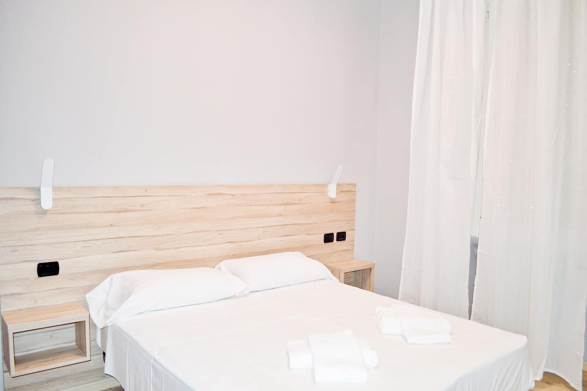 Una cama bien hecha con sábanas y almohadas blancas, un cabecero de madera, una mesita de noche y una cortina blanca.
