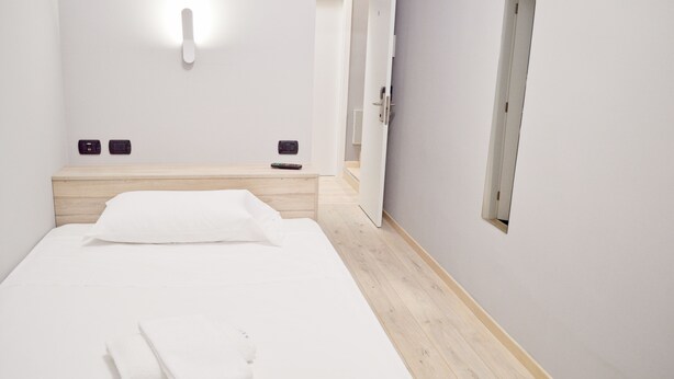 A minimalist hotel room with a wooden headboard, a single bed, a bedside table, and a wall-mounted light fixture. - Miu Hotel (Milan)