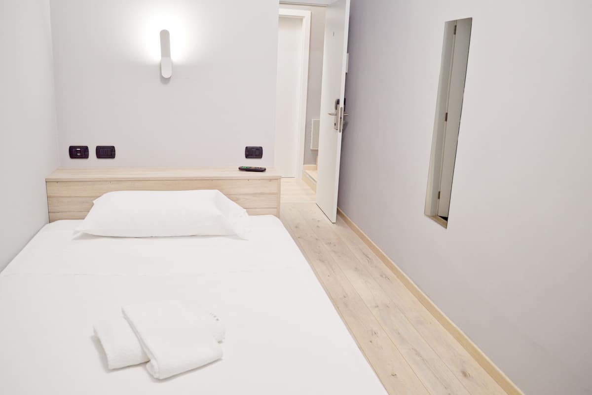 Habitación de hotel minimalista con cabecera de madera, una cama individual, una mesita de noche y un aplique de pared.