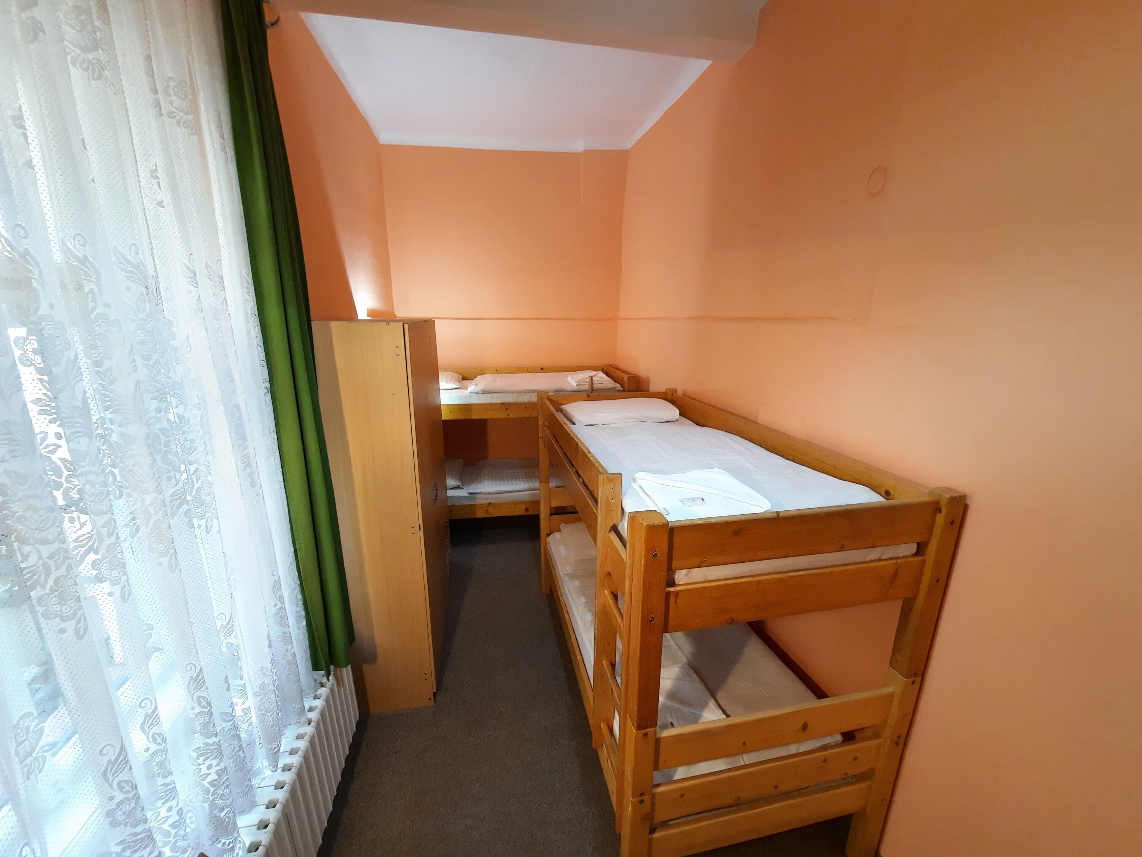 A dormitory room with bunk beds, a radiator, and a window with sheer curtains.
