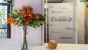 Un mostrador de recepción con un jarrón de flores naranjas y un cuenco de madera. La pared de fondo tiene un cartel de bienvenida en varios idiomas.